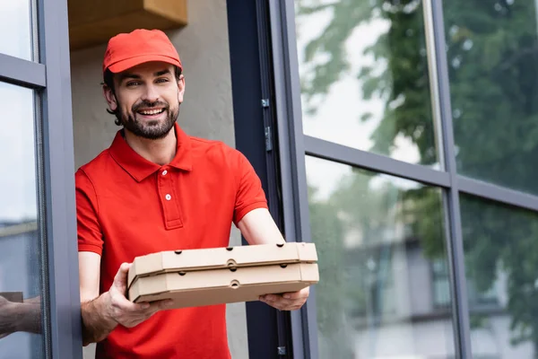 Bello cameriere guardando la macchina fotografica mentre tiene scatole di pizza vicino al caffè sulla strada urbana — Foto stock