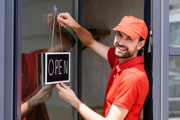 Vista lateral del camarero positivo en letrero colgante uniforme con letras abiertas en la puerta de la cafetería - foto de stock