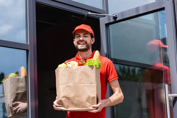 Courrier positif tenant sac en papier avec légumes frais et baguette dans la rue urbaine — Photo de stock