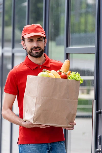 Mensajero sonriente sosteniendo bolsa de papel con frutas y verduras frescas cerca del edificio en la calle urbana - foto de stock