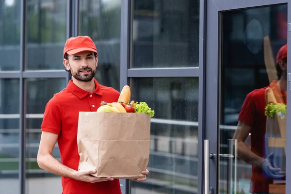 Guapo repartidor hombre sosteniendo bolsa de compras con frutas y verduras frescas en la calle urbana - foto de stock