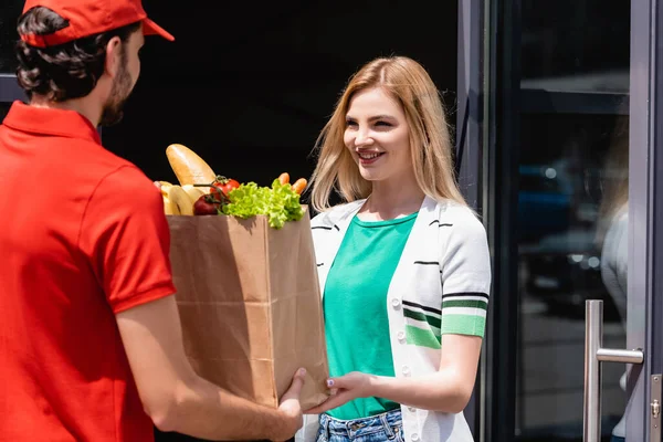 Enfoque selectivo de la mujer sonriente tomando bolsa de compras con verduras frescas de mensajero en la calle urbana - foto de stock