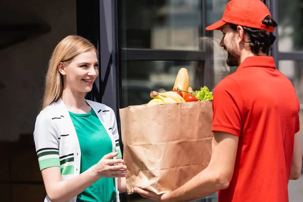Кур'єр тримає паперовий мішок з продуктовим магазином біля усміхненої жінки на міській вулиці — стокове фото