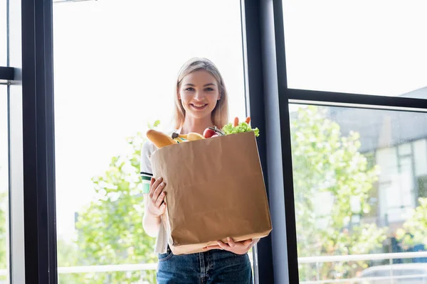 Mulher sorrindo segurando saco de compras com legumes frescos e olhando para a câmera em casa — Fotografia de Stock