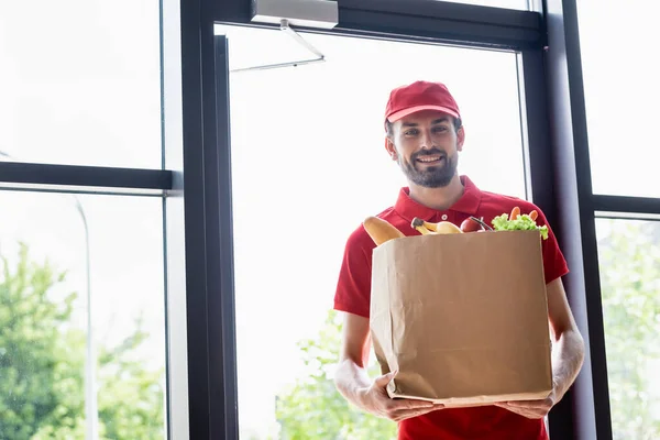 Mensajero sonriente sosteniendo la bolsa de la compra con el supermercado y mirando a la cámara - foto de stock