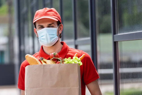 Courier in medical mask holding shopping bag with fresh vegetables on urban street — Stock Photo