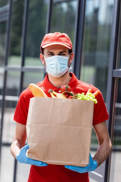 Hombre de reparto en máscara médica y guantes de látex sosteniendo bolsa de compras con comestibles en la calle urbana - foto de stock