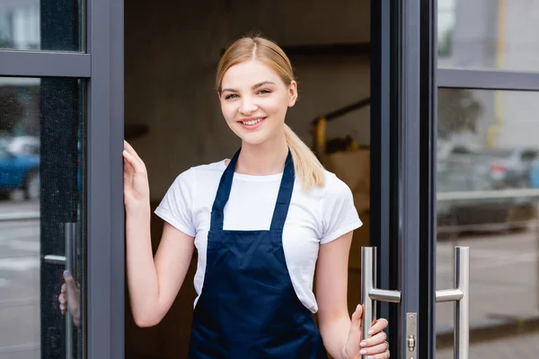 Cameriera sorridente guardando la fotocamera vicino alla porta aperta del caffè — Foto stock