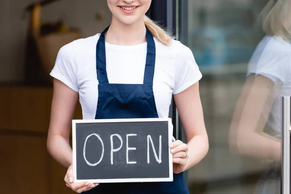 Cropped view of smiling waitress holding signboard with open lettering near cafe on urban street — Stock Photo