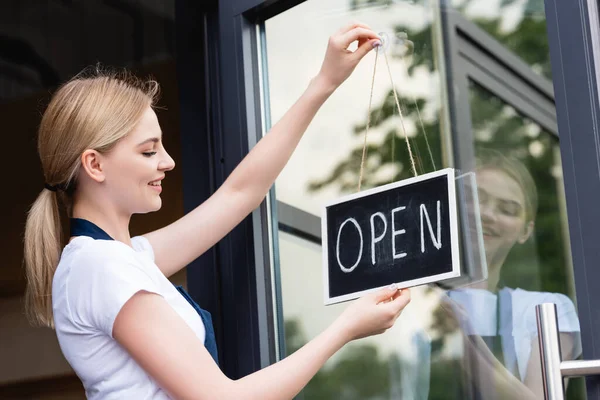 Side view of smiling waitress in apron hanging signboard with open lettering on door of cafe — Stock Photo