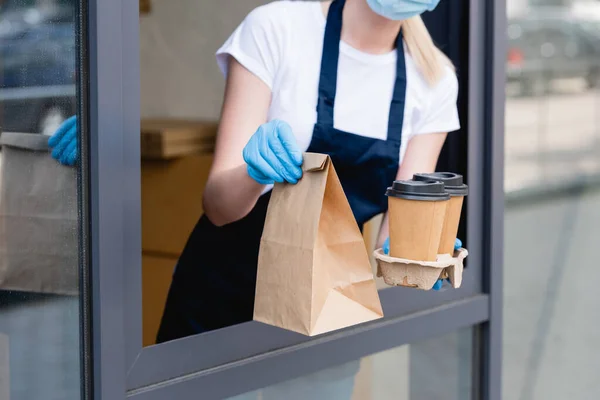 Vista recortada de la camarera en guantes de látex sosteniendo vasos de papel y paquete cerca de la ventana de la cafetería - foto de stock