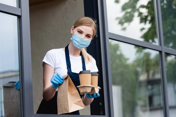 Vista de ángulo bajo de la camarera en guantes de látex y máscara médica sosteniendo vasos de papel y paquete cerca de la ventana de la cafetería - foto de stock