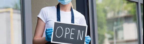 Panoramic shot of waitress in latex gloves and medical mask holding signboard with open word near window of cafe — Stock Photo