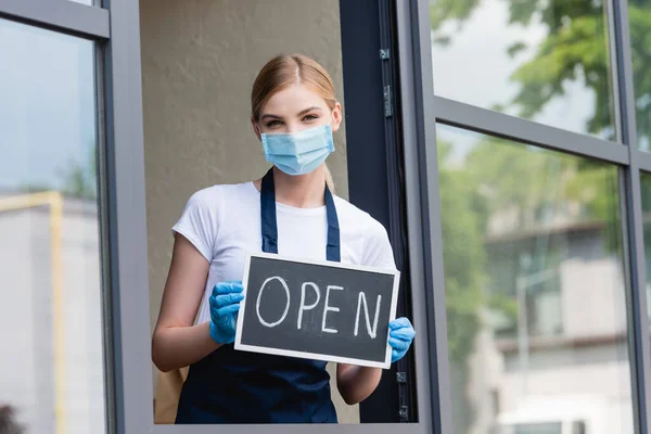 Waitress in latex gloves and medical mask holding signboard with open word near window of cafe — Stock Photo