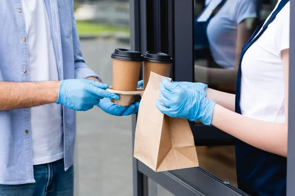 Vista cortada de homem segurando copos de papel e recebendo pacote de garçonete em luvas de látex perto de café — Fotografia de Stock