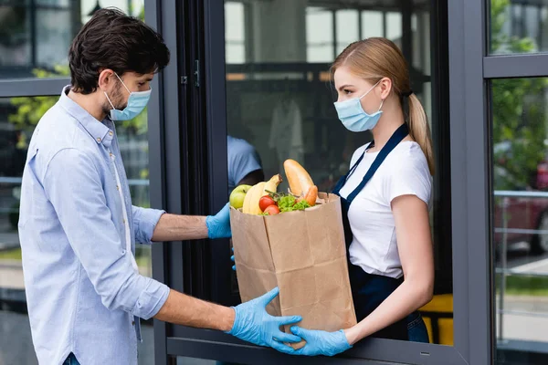 Side view of man taking shopping bag with fruits and vegetables from saleswoman near window — Stock Photo