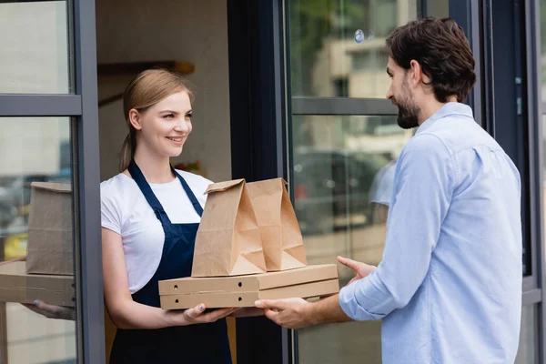 Atractiva camarera dando cajas de pizza y bolsas de papel al hombre en la entrada de la cafetería - foto de stock
