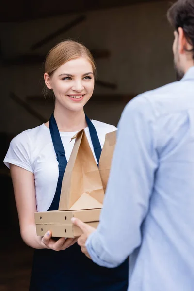 Selektiver Fokus der schönen Kellnerin, die den Mann anschaut und Pizzakartons und Papiertüten am Eingang des Gebäudes verteilt — Stockfoto