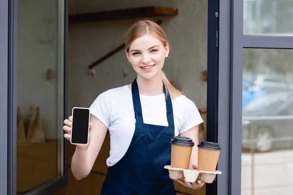 Front view of beautiful waitress holding smartphone and paper cups near window of cafe — Stock Photo