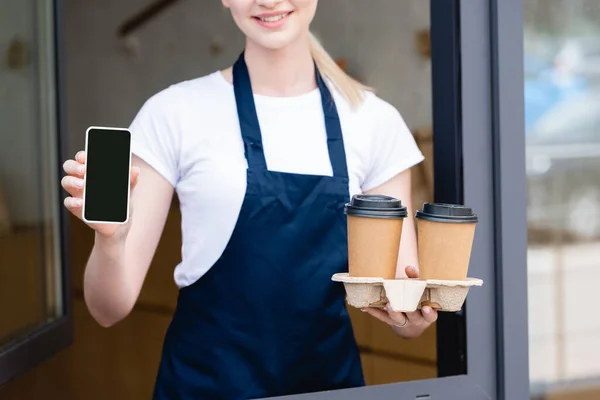 Обрезанный вид на баристу с бумажными чашками и смартфоном рядом с кафе — стоковое фото