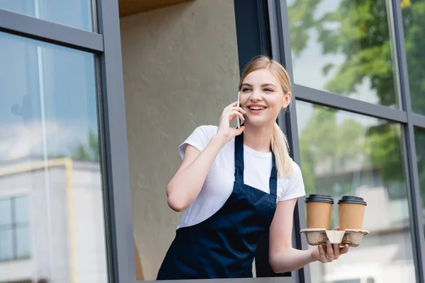 Vista de ángulo bajo de camarera sonriente hablando en el teléfono inteligente y sosteniendo vasos de papel cerca de la ventana de la cafetería - foto de stock