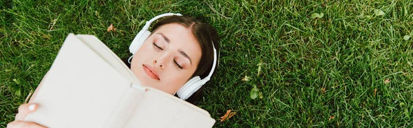 Panoramic shot of girl in wireless headphones reading book and lying on green grass — Stock Photo