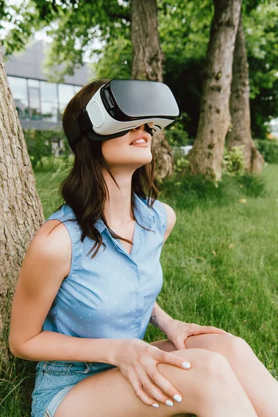 Щаслива молода жінка в гарнітурі віртуальної реальності, що сидить біля зелених дерев — стокове фото
