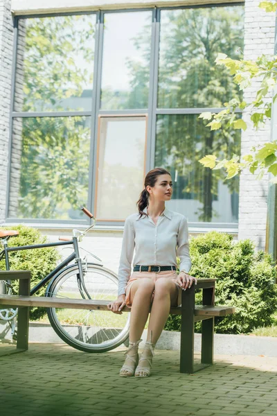 Attraktive Geschäftsfrau sitzt auf Bank neben Fahrrad und Gebäude — Stockfoto