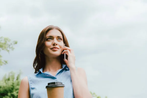 Low angle view of businesswoman holding disposable cup and talking on smartphone against cloudy sky — Stock Photo