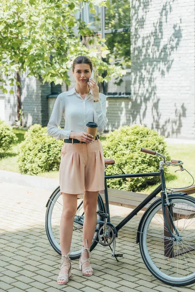 Atractiva mujer de negocios hablando en el teléfono inteligente y sosteniendo la taza de papel mientras está cerca de la bicicleta - foto de stock