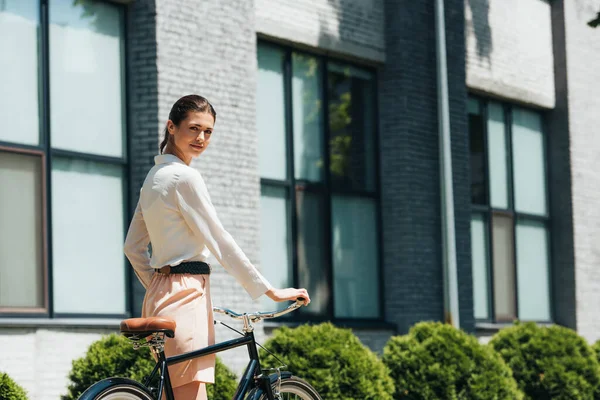 Atractiva mujer de negocios caminando con bicicleta cerca de un edificio moderno - foto de stock