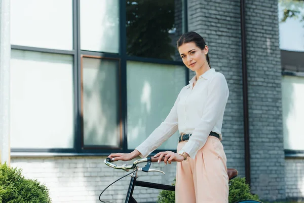 Bela jovem empresária de pé com bicicleta perto do edifício moderno — Fotografia de Stock