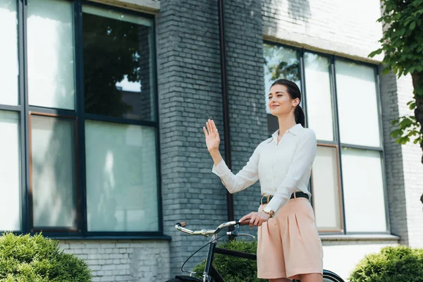 Joyeuse jeune femme d'affaires debout avec vélo et main agitant — Photo de stock