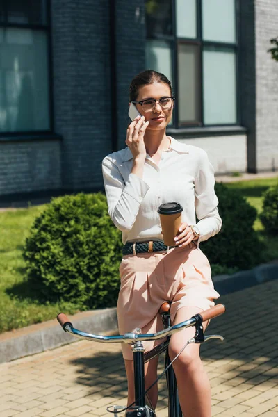 Atractiva mujer de negocios en gafas hablando en el teléfono inteligente y sosteniendo taza de papel cerca de la bicicleta - foto de stock