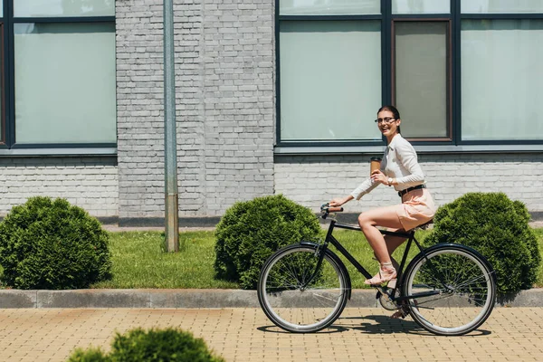 Selective focus of cheerful businesswoman in glasses riding bicycle and holding paper cup — Stock Photo