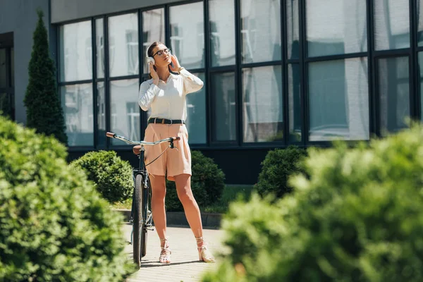 Selektiver Fokus der attraktiven Geschäftsfrau mit Brille, die Musik hört und drahtlose Kopfhörer berührt, während sie in der Nähe des Fahrrads steht — Stockfoto