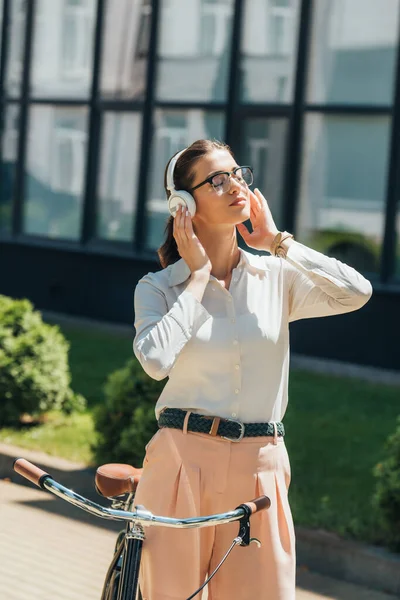 Young businesswoman in glasses listening music and touching wireless headphones while standing near bicycle — Stock Photo