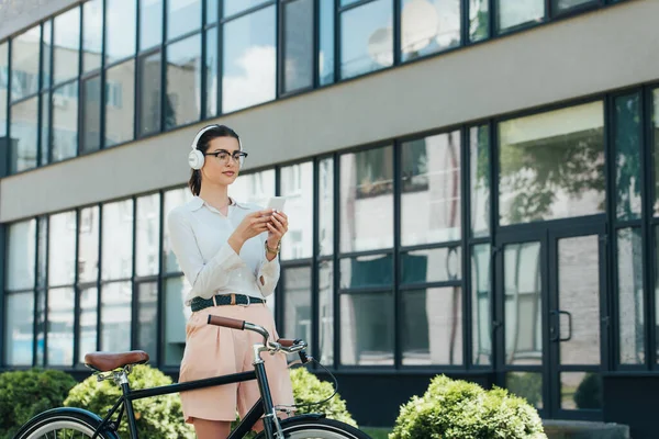 Atractiva mujer de negocios en gafas escuchando música en auriculares inalámbricos y utilizando un teléfono inteligente cerca de la bicicleta - foto de stock