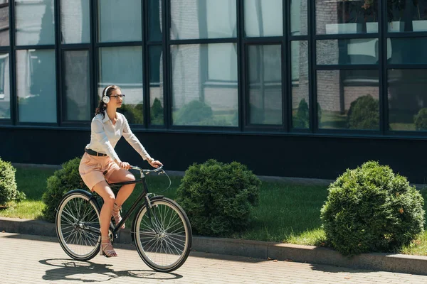 Bela empresária em óculos ouvir música em fones de ouvido sem fio e andar de bicicleta perto do edifício — Fotografia de Stock