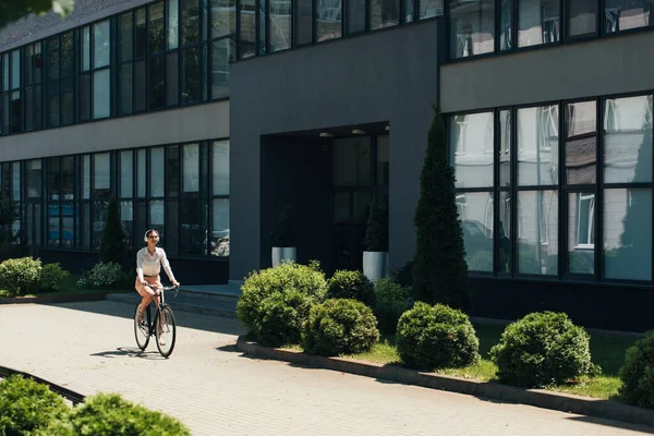 Молодая бизнесвумен в очках слушает музыку в беспроводных наушниках и катается на велосипеде возле здания — стоковое фото