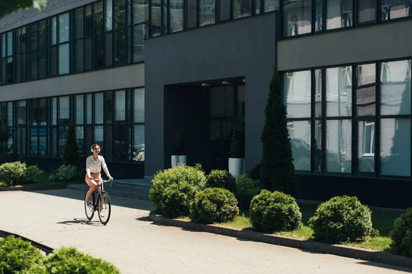 Joven mujer de negocios en gafas escuchando música en auriculares inalámbricos y montar en bicicleta cerca del edificio - foto de stock