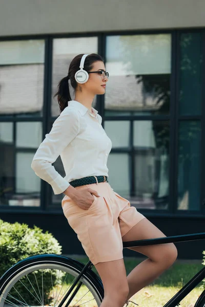 Vista lateral de hermosa mujer de negocios con la mano en el bolsillo escuchando música en auriculares inalámbricos y bicicleta de montar cerca del edificio - foto de stock