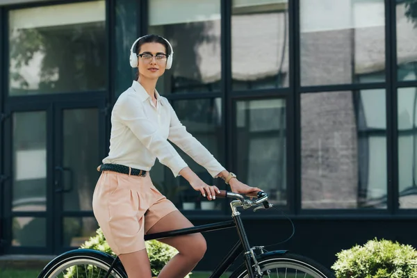 Schöne Geschäftsfrau mit Brille, die Musik in drahtlosen Kopfhörern hört und in der Nähe von Gebäuden Fahrrad fährt — Stockfoto