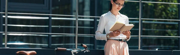 Panoramic concept of businesswoman in glasses reading book near bicycle and modern building — Stock Photo