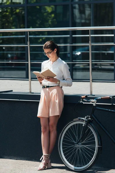 Бизнесвумен в очках читает книгу возле велосипеда и современного здания — стоковое фото