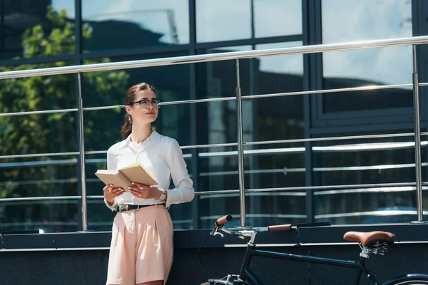 Beautiful businesswoman in glasses holding book near bicycle and modern building — Stock Photo