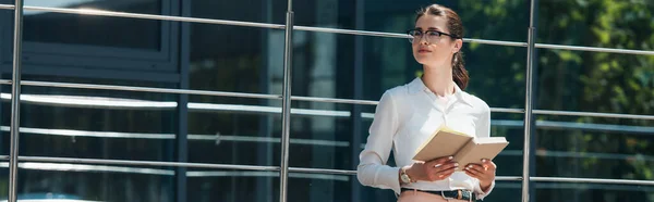 Culture panoramique de femme d'affaires dans des lunettes tenant livre près du bâtiment moderne — Photo de stock