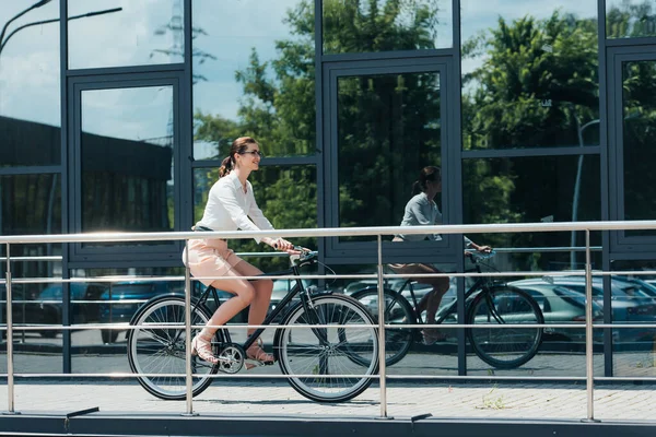 Joyeuse jeune femme d'affaires à lunettes équitation vélo près du bâtiment — Photo de stock