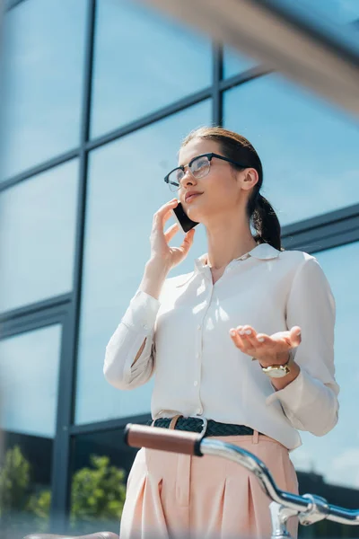 Selective focus of businesswoman in glasses talking on smartphone near building and bicycle — Stock Photo