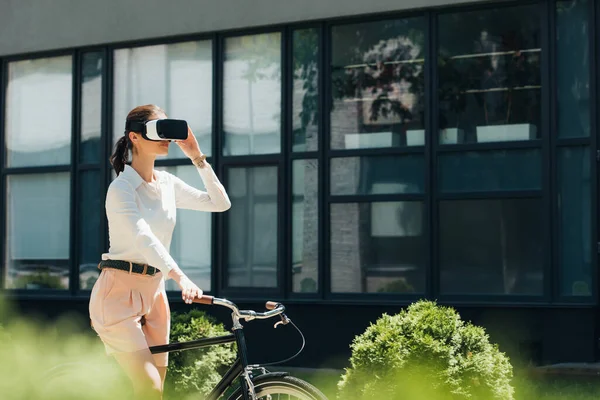 Messa a fuoco selettiva della donna d'affari in realtà virtuale auricolare in bicicletta vicino alla costruzione — Foto stock